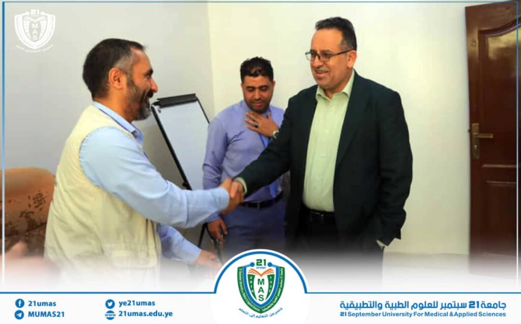 Two men shake hands in a university office as a colleague observes, with the university crest and banner at the bottom of the frame.