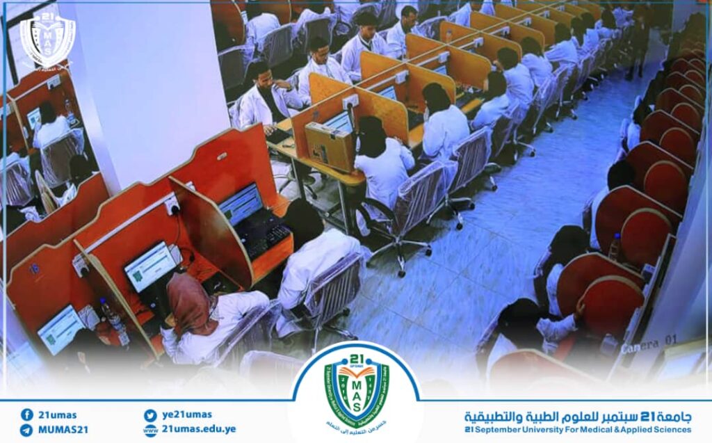 Panoramic view of students in white coats working at computer desks with orange dividers in a university lab; logo and contact info at the bottom.