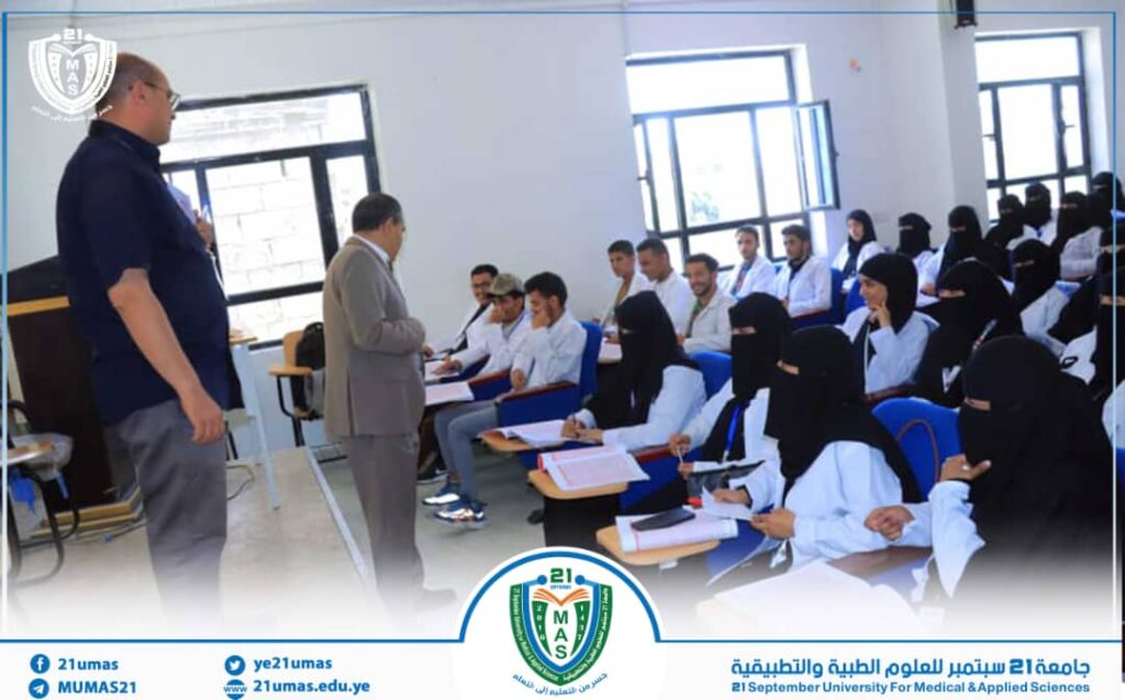 University lecture in progress: two men speak at the front of a classroom while students, many wearing hijabs, take notes and listen attentively.