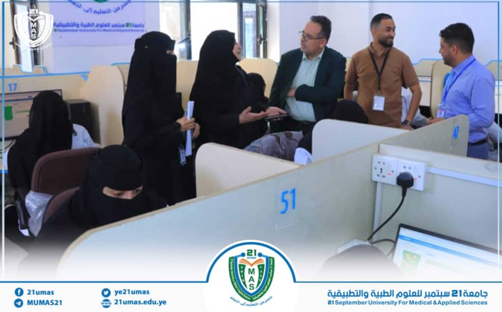 Group of staff and students, including veiled women, talking beside numbered cubicles in a university service area with the 21MAS banner/logo visible.