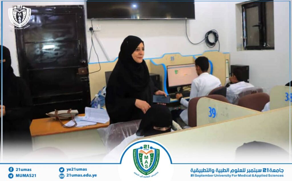Women in black abayas work and study in a computer lab, with a instructor standing beside student desks and monitors on the wall-mounted setup in a university setting.