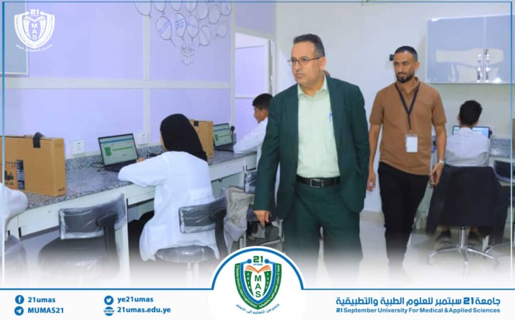 University lab scene: staff in white coats at computer stations while an administrator in a green suit walks through the room, logo visible in background and foreground.