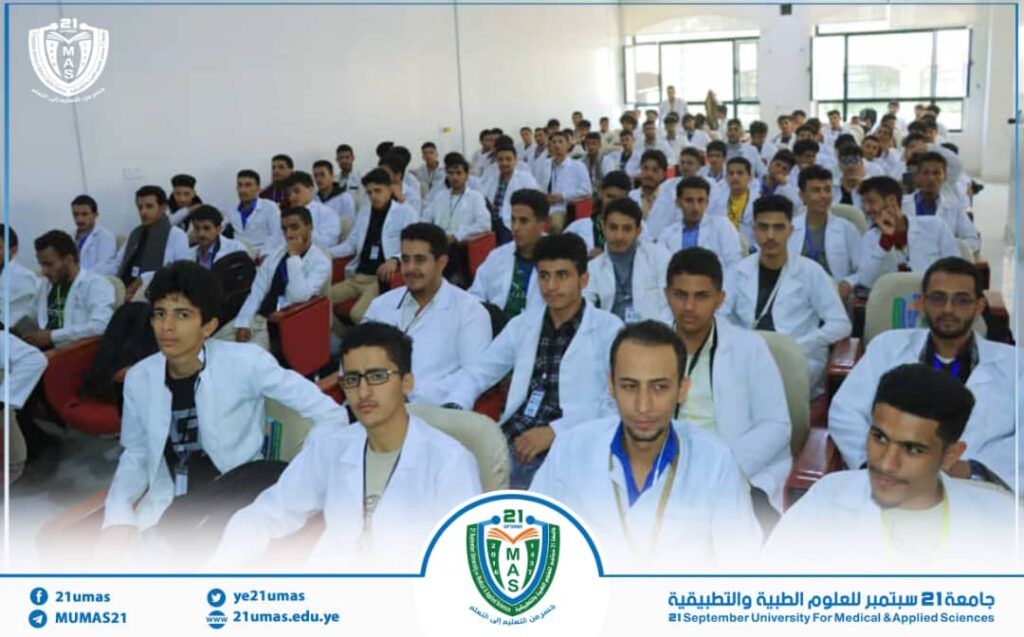 Group of students in white lab coats seated in a bright lecture hall, wearing lanyards; a university banner and logo are visible at the front.