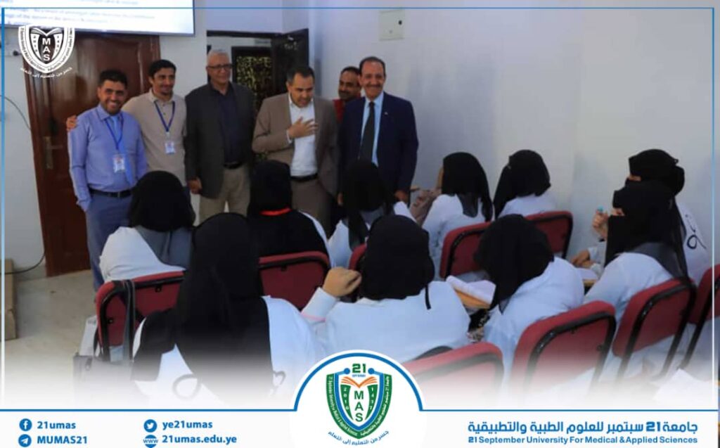 Group of male speakers address female medical students in white coats in a lecture hall, with the university emblem visible on the bottom edge.