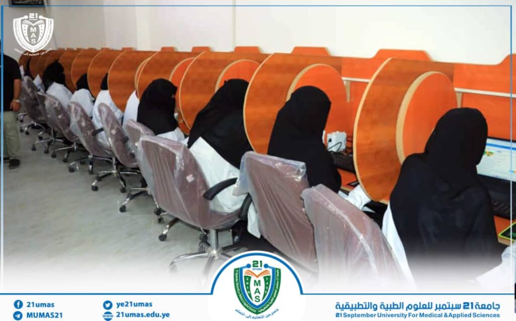 Women in black abayas sit at individual computer desks in a university computer lab, with orange privacy dividers between stations.