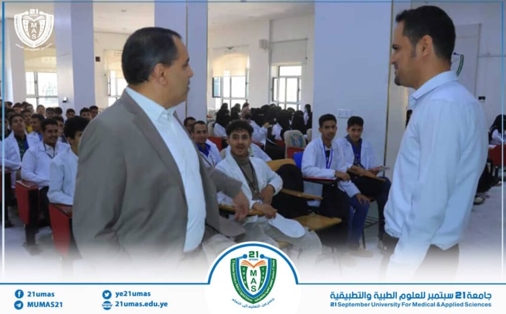 Two men stand at the front of a university lecture hall, addressing a roomfull of seated students with university branding visible.