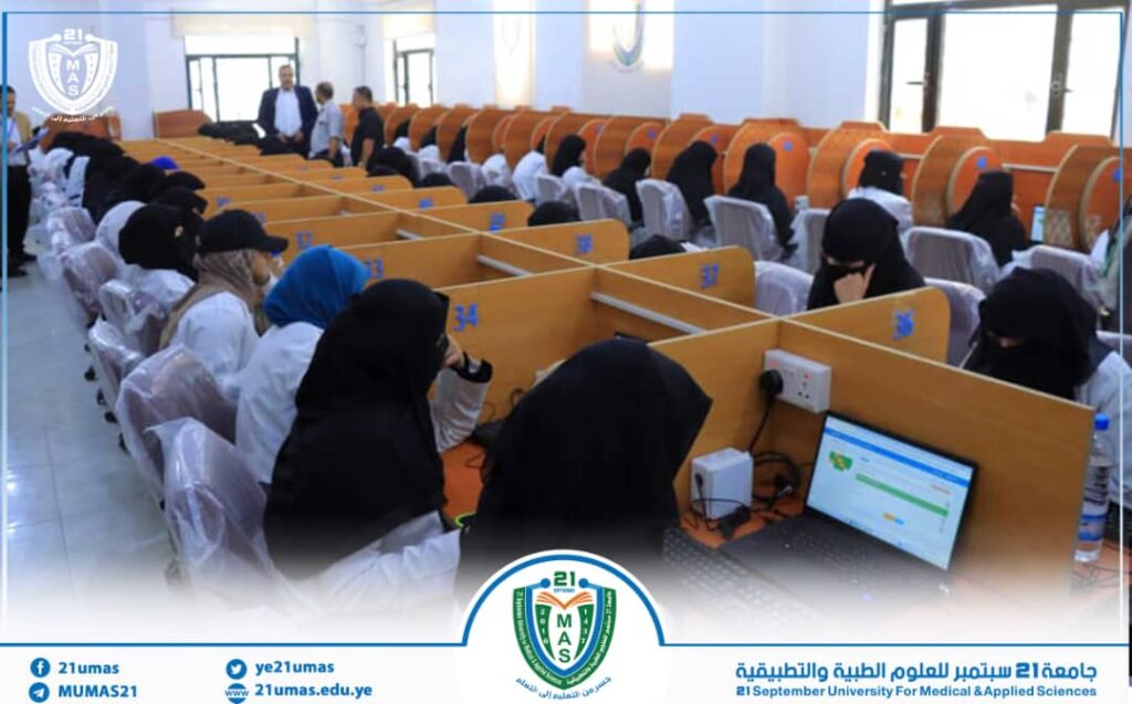 Students in white coats and hijabs sit at computer desks in a large lab with orange dividers and a university emblem on the wall/banner in the background.