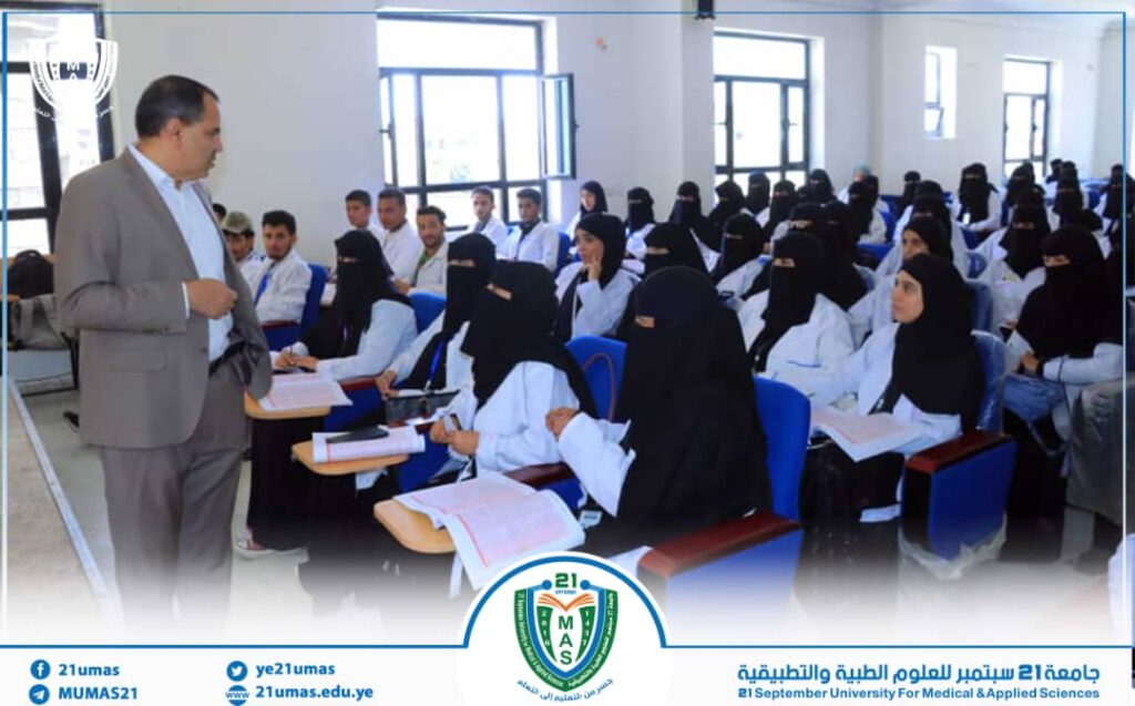A male lecturer in a gray suit addresses a classroom full of female students wearing black hijabs in a university lecture hall.