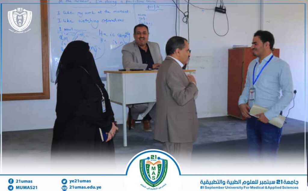 Man at a podium in a classroom speaks to a man in a light blue shirt; a veiled woman and another man stand nearby as notes are on the whiteboard behind them.