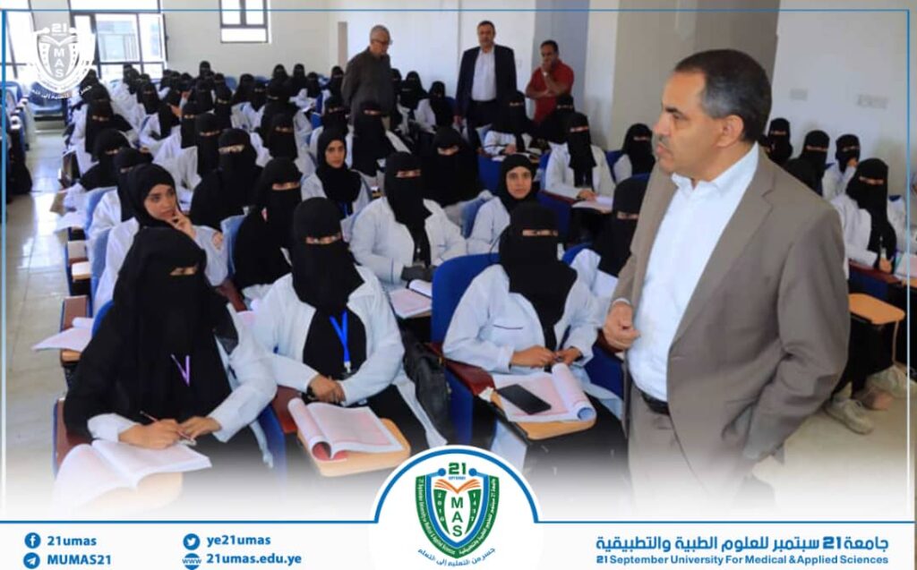 Male instructor in a beige suit stands at the front of a classroom addressing a large group of female students in white coats and black hijabs; students take notes as the logo appears at the bottom.