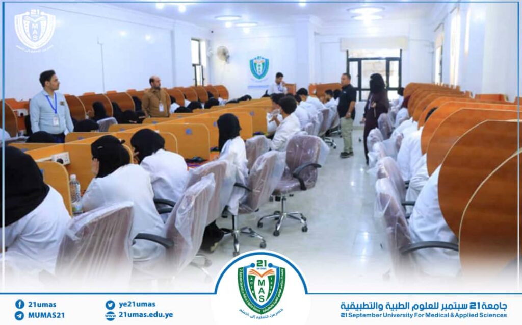 Lecture hall at 21 September University for Medical & Applied Sciences; students in white coats sit at curved wooden desks, staff at front.