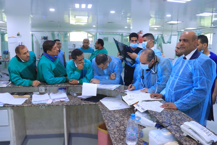 Medical staff in green and blue scrubs, some wearing masks, gather around a hospital counter reviewing documents.