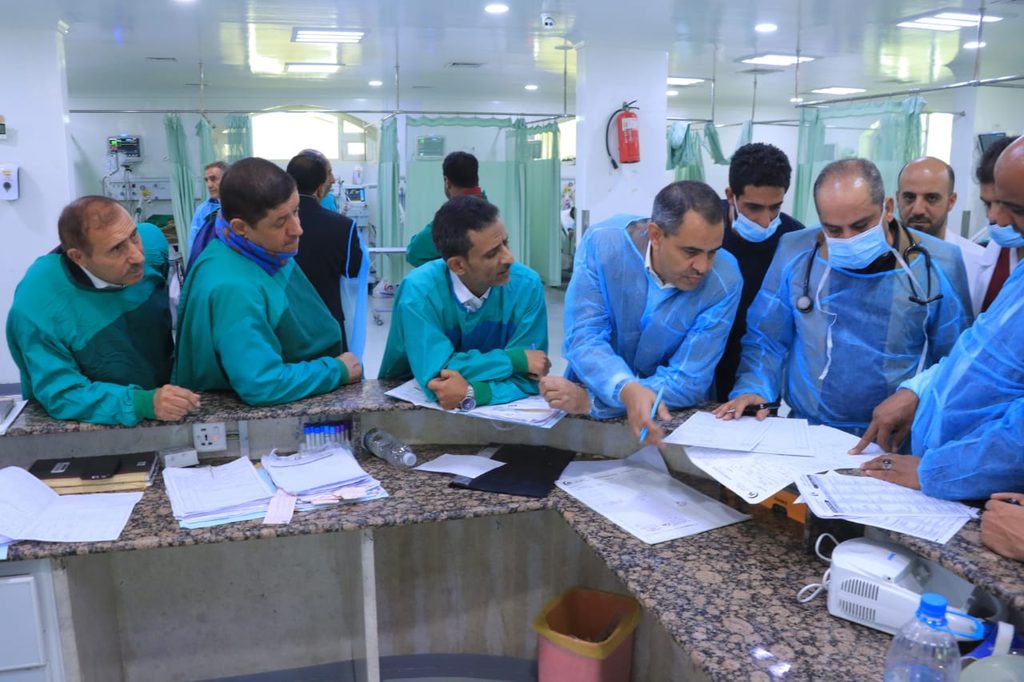 Medical team in green and blue gowns gathered around a clinical counter, reviewing papers and patient charts in a hospital bay.