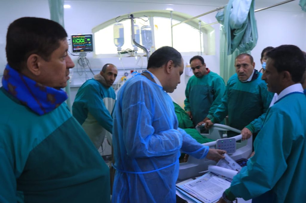 Medical team in green scrubs gathers around a patient on a hospital bed with monitors and IVs nearby (ICU/operating room).