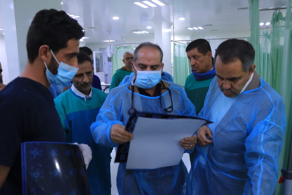Medical team in blue protective gowns and masks review a clipboard in a bright hospital ward.