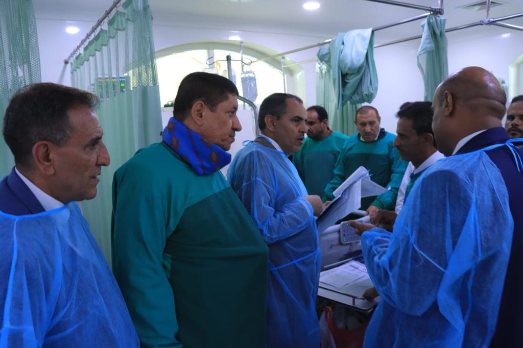 Medical staff in green and blue gowns gathered around a patient monitoring station in a ward with green curtains closed in the background