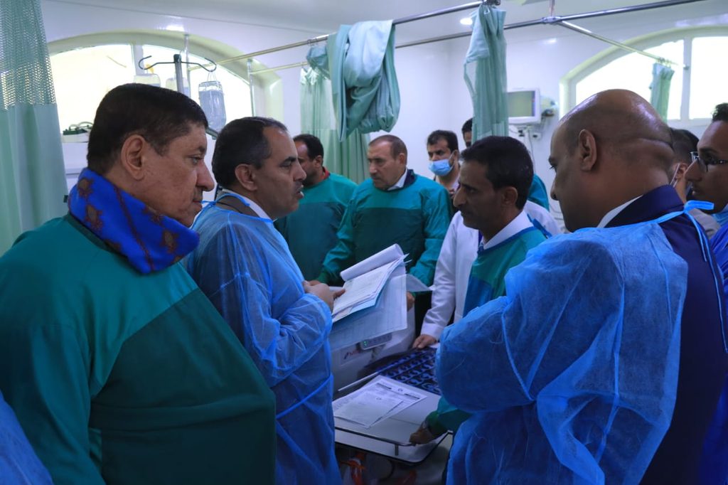 Medical team in teal gowns stands around a patient monitor on a hospital ward table.