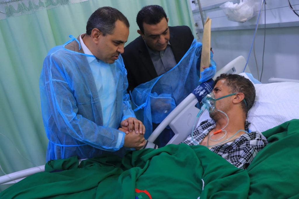 Two men in blue protective gowns hold the hand of a patient in a hospital bed wearing an oxygen mask, showing support.