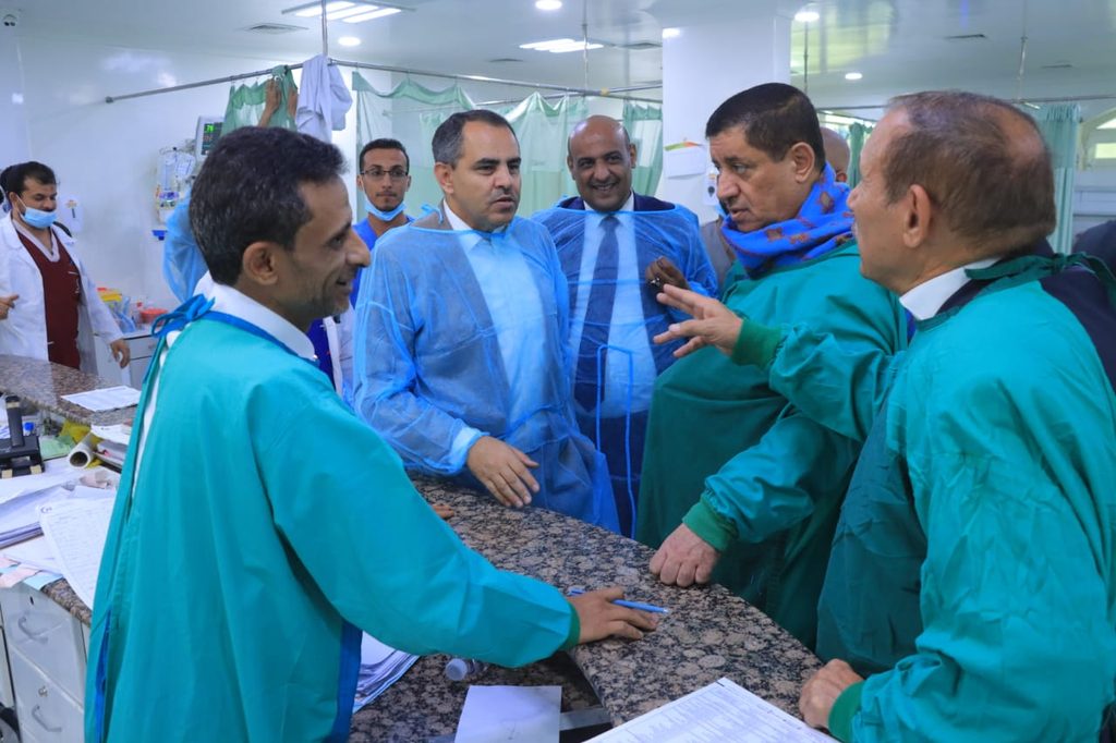 Group of men in disposable blue and green surgical gowns gather around a marble hospital counter, discussing in a ward with green curtains.