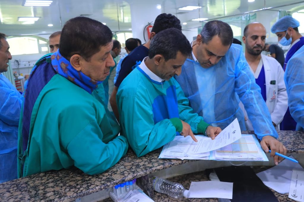 Group of medical staff in green scrubs and blue gowns review documents on a hospital counter, focused on a form.