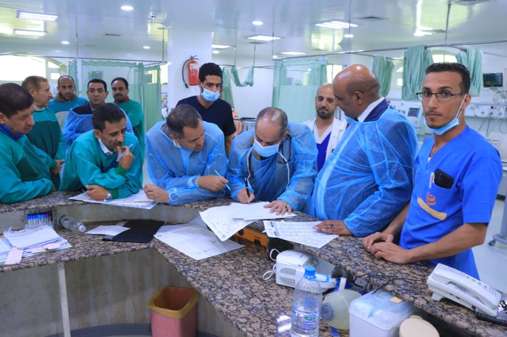 Medical team gathers around a hospital counter, signing and reviewing forms, some wearing masks and blue gowns.