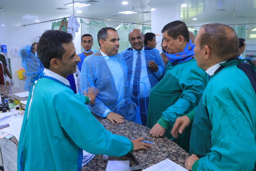 Hospital staff and visitors in protective gowns stand at a check-in counter, discussing paperwork in a clinical setting.