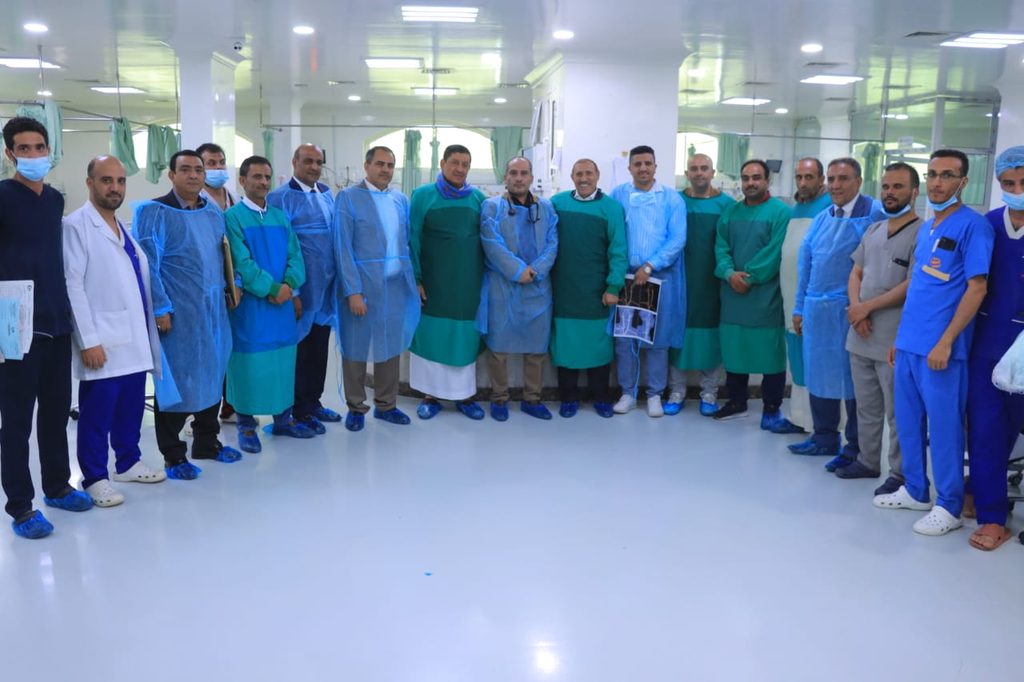 Group of doctors and nurses in blue and green gowns posing for a group photo in a bright hospital ward. They stand in a semicircle on a clean white floor, some wearing masks and protective gear.