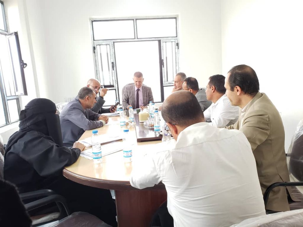 Group of professionals seated around a rectangular table in a bright meeting room, discussing with papers and water bottles on the table.