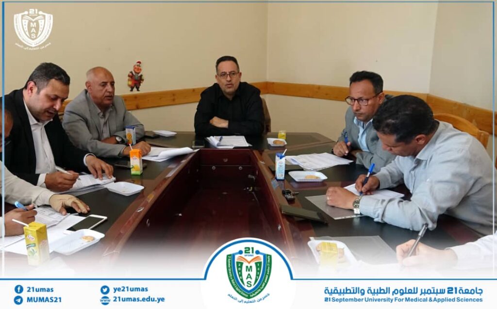 Six men sit around a conference table, reviewing and signing documents in a formal meeting room with a university crest visible on the table border.