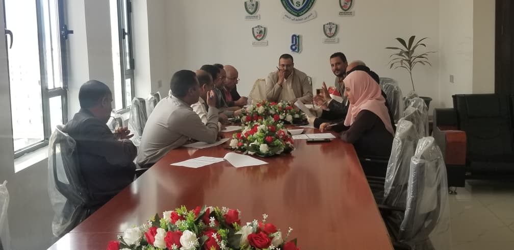 A diverse group of professionals sit around a long conference table in a meeting room, engaged in discussion with papers and a central floral arrangement in front of them.