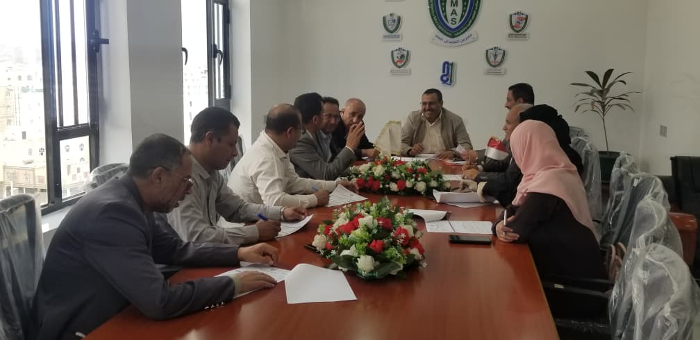 A group of professionals seated around a long conference table discussing documents in a bright office with flower arrangements center stage.