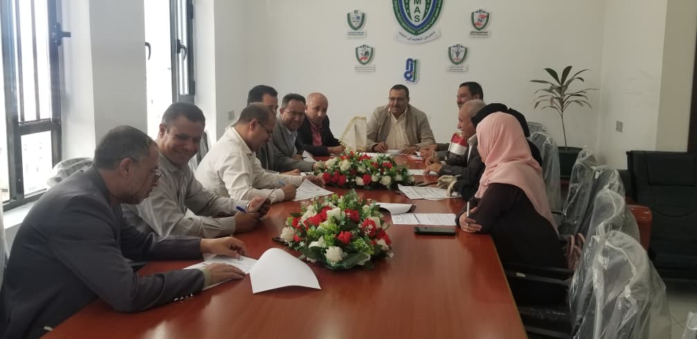 Group of professionals seated around a long conference table reviewing documents with a flower centerpiece in a bright office meeting room.