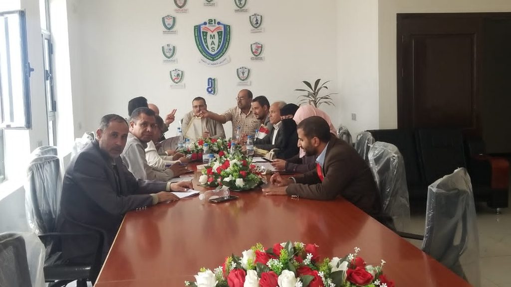 Group of men in business attire seated around a long conference table in discussion, with a red-and-white floral centerpiece and wall logos in the background.