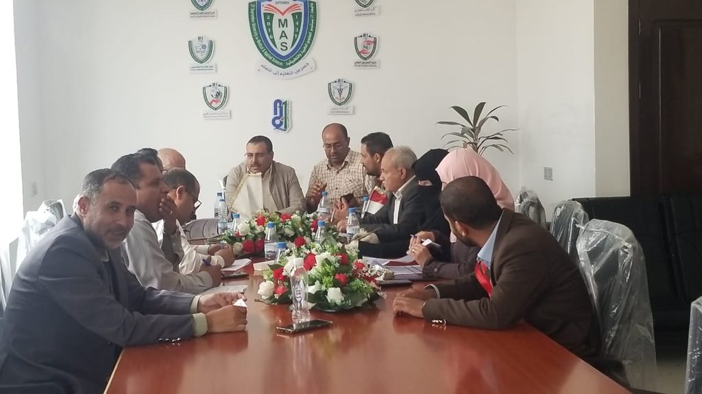 Group of professionals in a formal meeting around a long conference table with a floral centerpiece and bottled water.