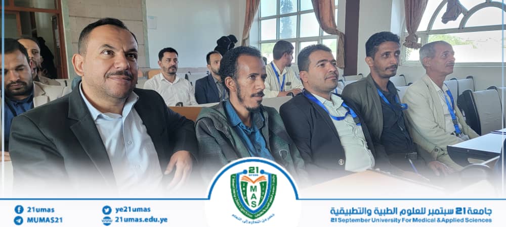 Group of men in a conference room seated in a row, wearing lanyards, watching a presentation; a university-branded banner with a crest and social links is visible at the front.