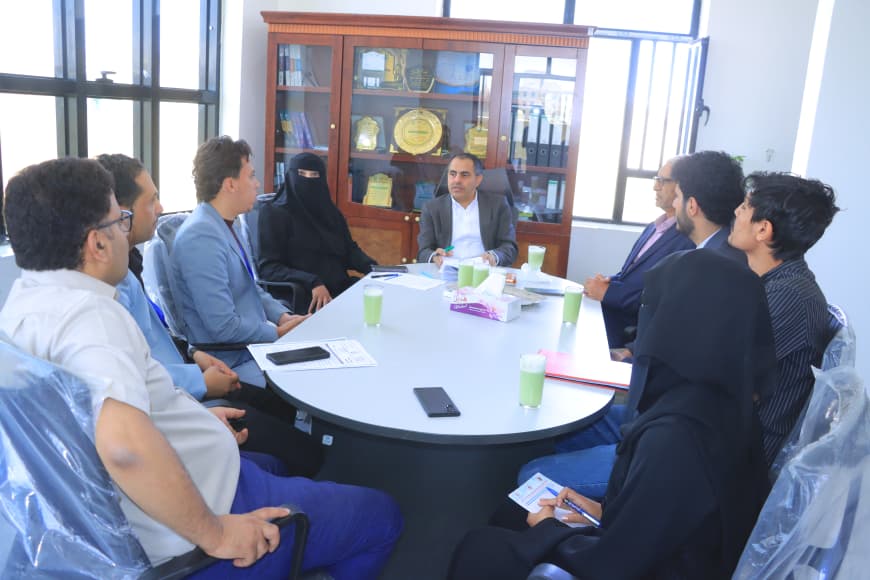 Team meeting around a round table in a bright office; a man at the head speaks as colleagues listen, with a trophy cabinet in the background.