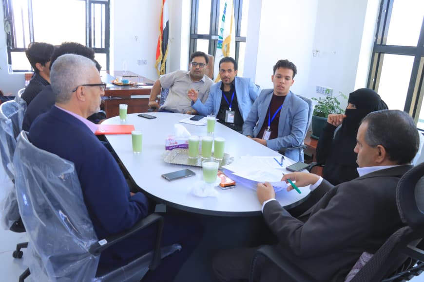 Eight people sit around a round conference table in a bright meeting room, discussing documents with lanyards visible on several attendees.
