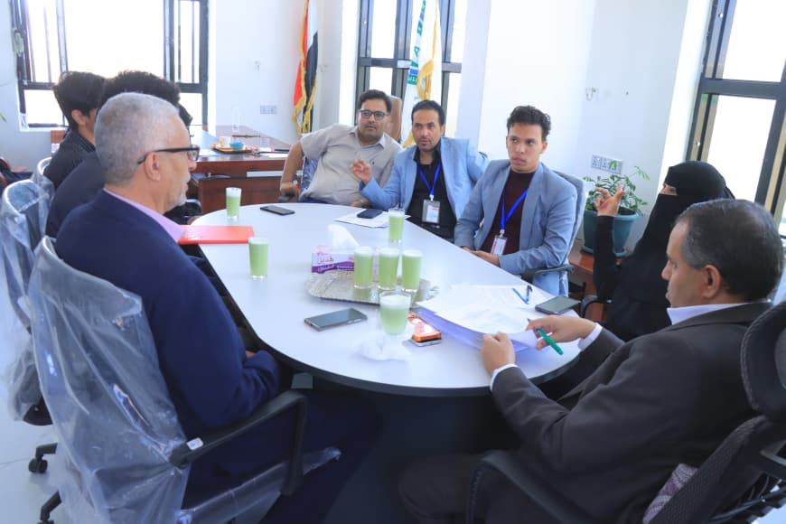 Group of eight people around a conference table in a bright office, discussing with papers and green beverages on the table.