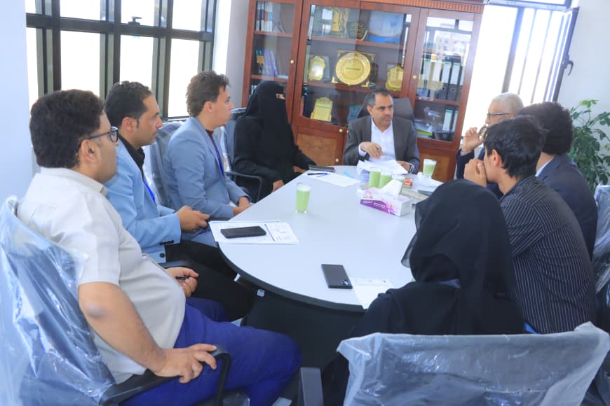 Diverse group of professionals seated around an oval conference table in a meeting, with papers, phones, and cups on the table; trophy cabinet in the background.