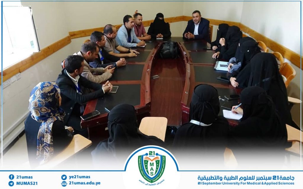 Group of men and women in a meeting around a curved conference table; a man at the far end speaks to the group.