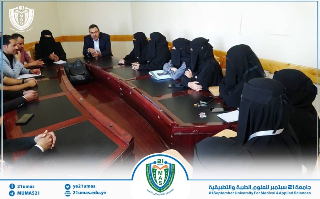 A formal meeting around a long conference table with a man in a suit speaking, flanked by women in black hijabs on the right; university logo appears in the corner and a blue banner at the bottom.