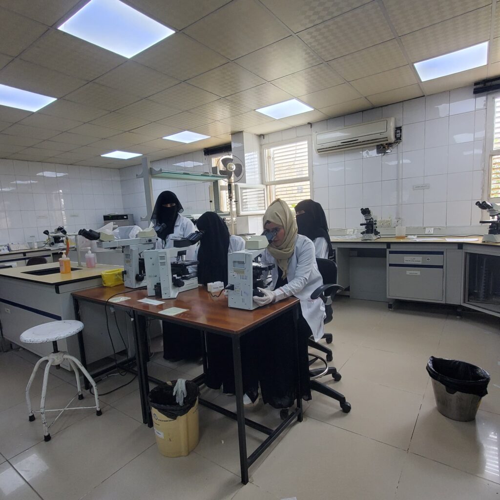 Laboratory scene with women in white coats using microscopes at a long bench; tiled walls and ceiling lights.