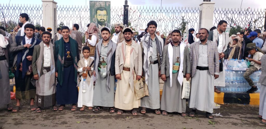 Group of men and a boy standing in a row outdoors, wearing traditional robes and scarves, near a metal fence with a portrait in the background.