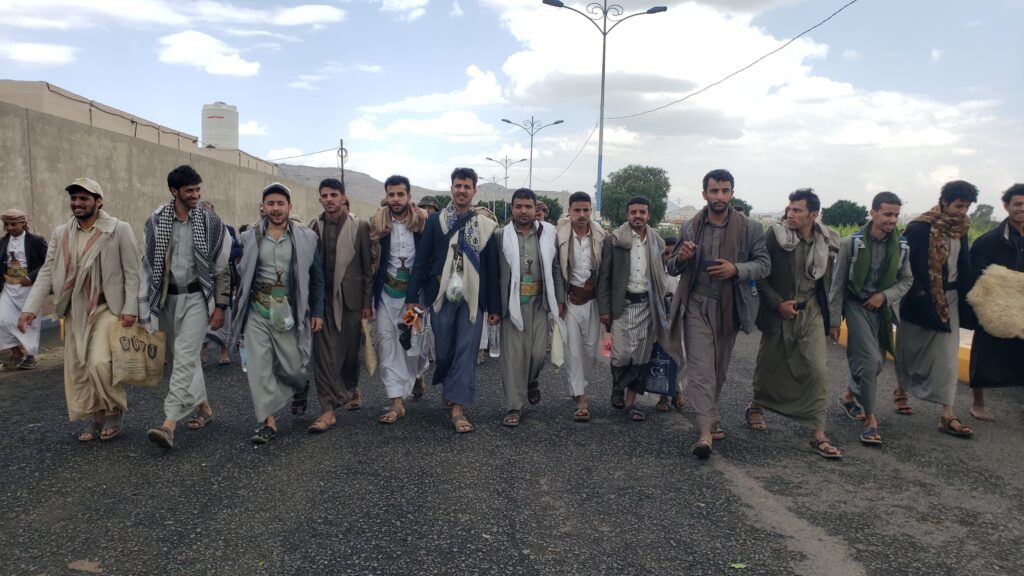 Group of men in traditional clothing walking together down a paved road on a sunny day in a line formation along a wall.