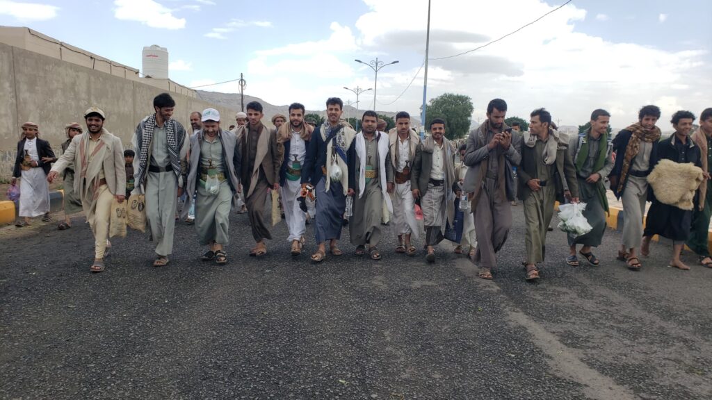 A line of men walking down a paved street in traditional clothing, many carrying bags or water bottles.