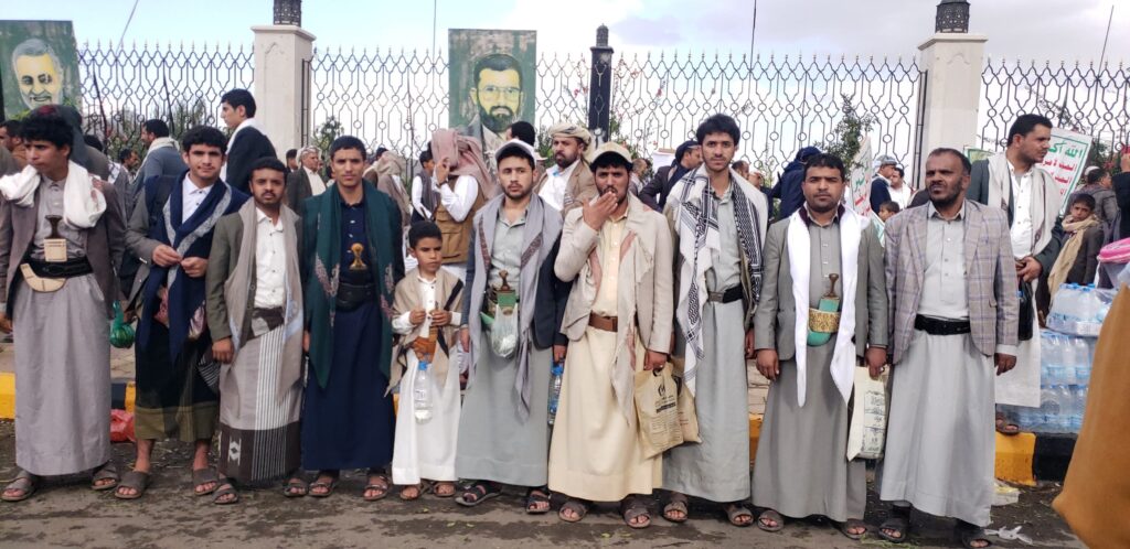 Group of men (and a boy) in traditional Yemeni attire standing in a line at an outdoor event near a decorative fence with portraits in the background, holding bags/water bottles and wearing sandals.