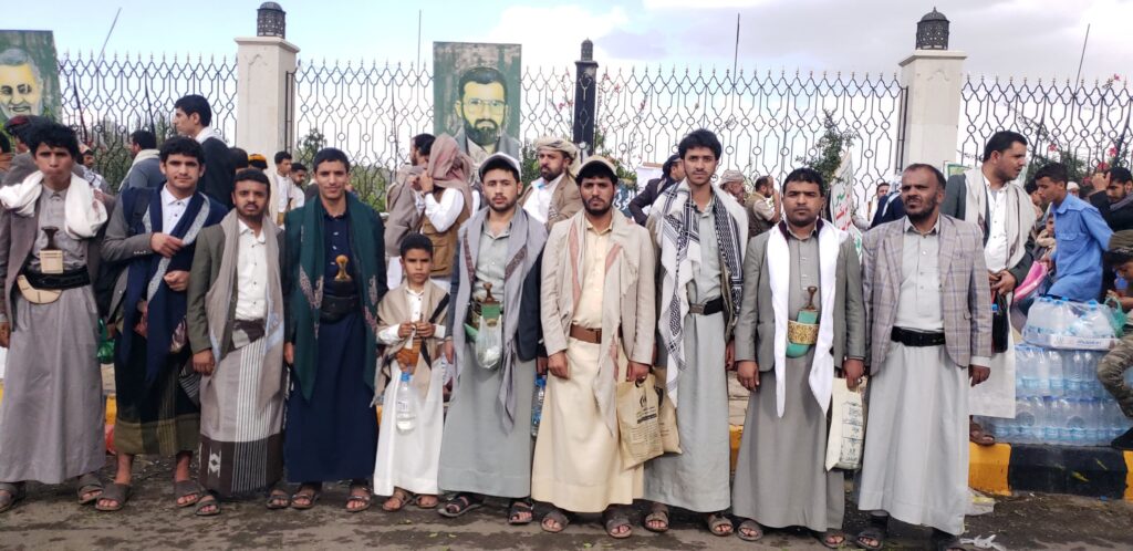 Group of men wearing traditional robes and scarves standing in a line outdoors, with a metal fence and posters visible behind them.
