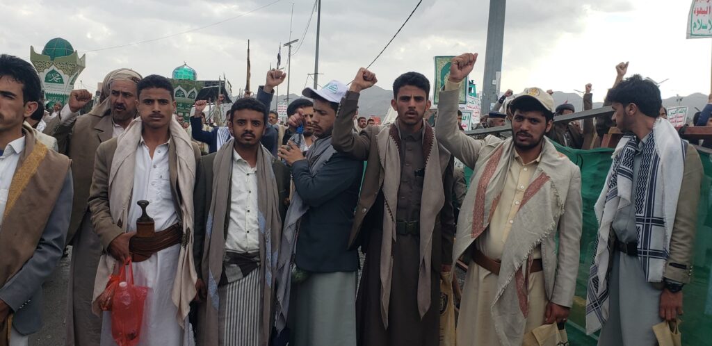 Group of men in traditional robes at a protest, raising fists with banners and signs in Arabic behind them.