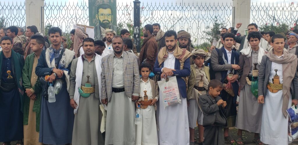 Group of men and boys in traditional Yemeni robes with daggers in belts, gathered in front of a wrought-iron fence and a large portrait mural.