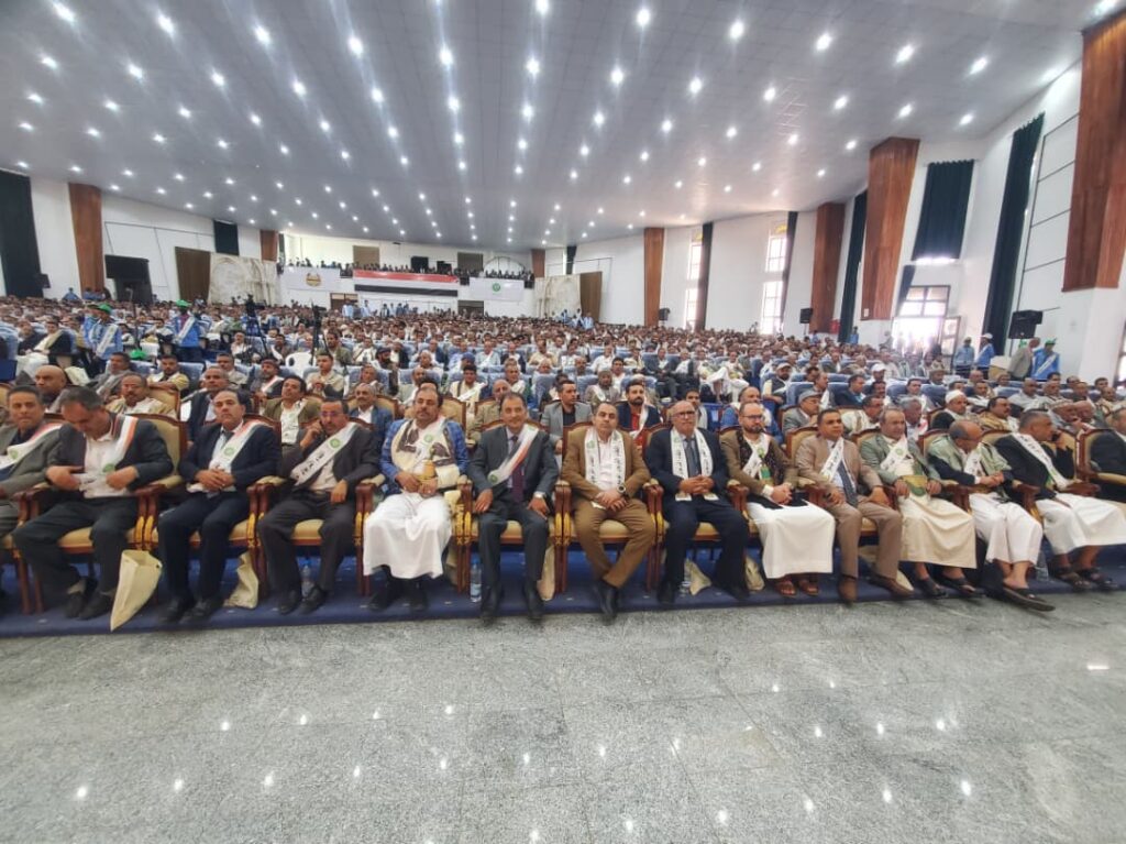 IMG-20260919-WA0053 - جامعة 21 سبتمبر Large conference hall filled with hundreds of attendees; front row in formal dress with sashes, facing the stage.